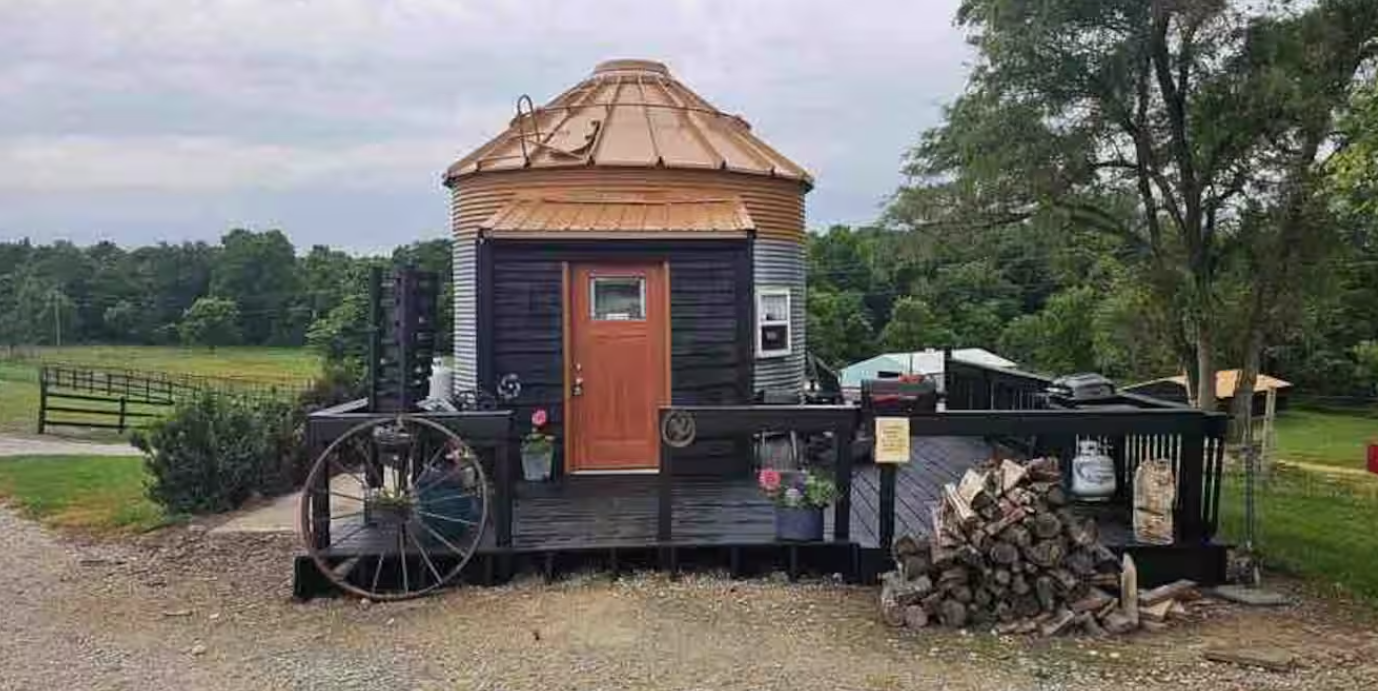 A small, round house made from a converted metal grain silo sits on a black wooden deck with potted plants, a large wagon wheel, firewood, and rural scenery—one of the coolest Airbnbs for a unique countryside escape.