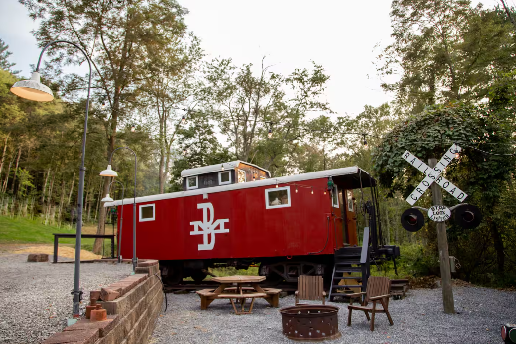 A bright red train caboose converted into a cabin sits among trees, with outdoor seating, a fire pit, and a railroad crossing sign nearby—one of the coolest Airbnbs for a rustic, unique lodging experience.