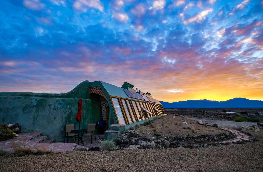 A unique, earth-sheltered greenhouse with large slanted windows sits in a rocky desert landscape at sunrise—one of the coolest Airbnbs, framed by vibrant clouds and distant mountains in the background.