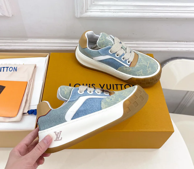 A pair of blue and beige Louis Vuitton sneakers with white laces and gum soles are displayed on an orange Louis Vuitton box, perfect for elevating your clothes collection. A hand holds one shoe, with a branded dust bag partially visible beside the box.