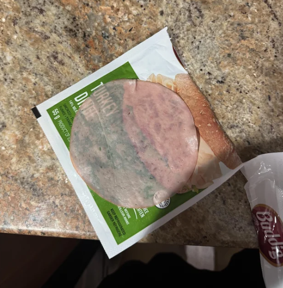 A slice of turkey deli meat is partially outside of its packaging, lying on a speckled kitchen countertop. The turkey slice is thin and slightly translucent, revealing the label underneath.