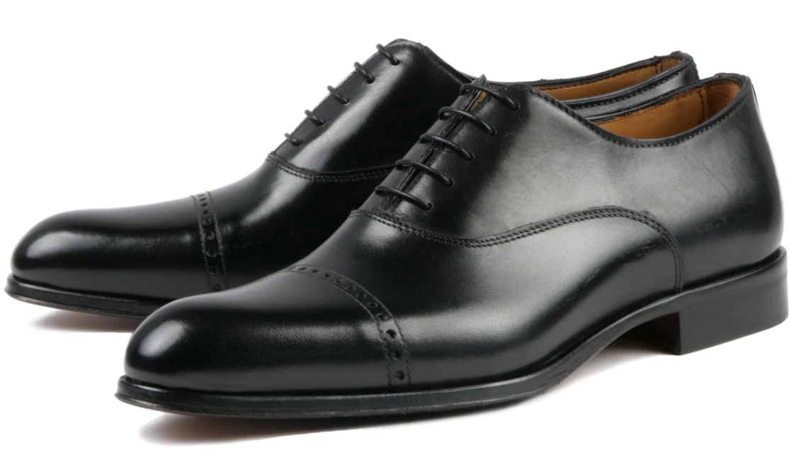 A pair of classic black leather Oxford dress shoes with closed lacing, low heels, and a slightly rounded toe—an essential addition to formal clothes, featuring a cap-toe design with subtle decorative perforations.