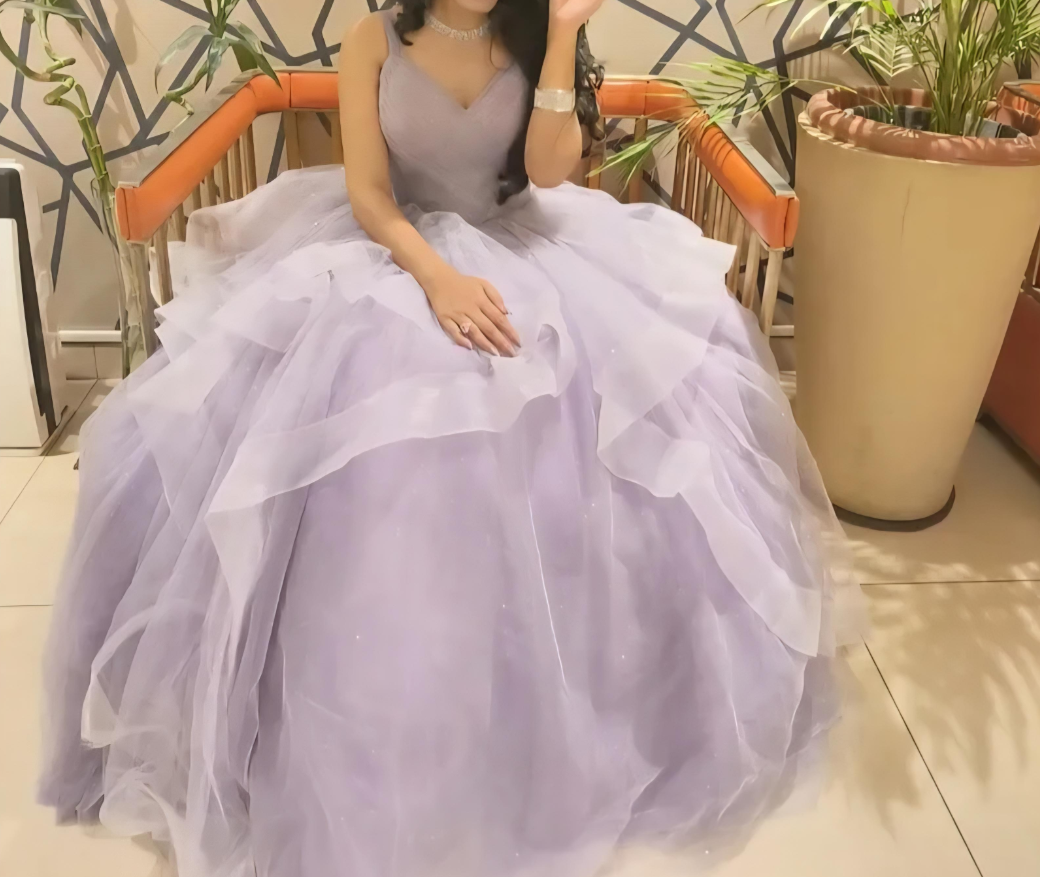 A person wearing voluminous, light purple clothes—a layered tulle ball gown—sits on an orange cushioned bench, surrounded by large potted plants indoors. Their face is partially out of frame.