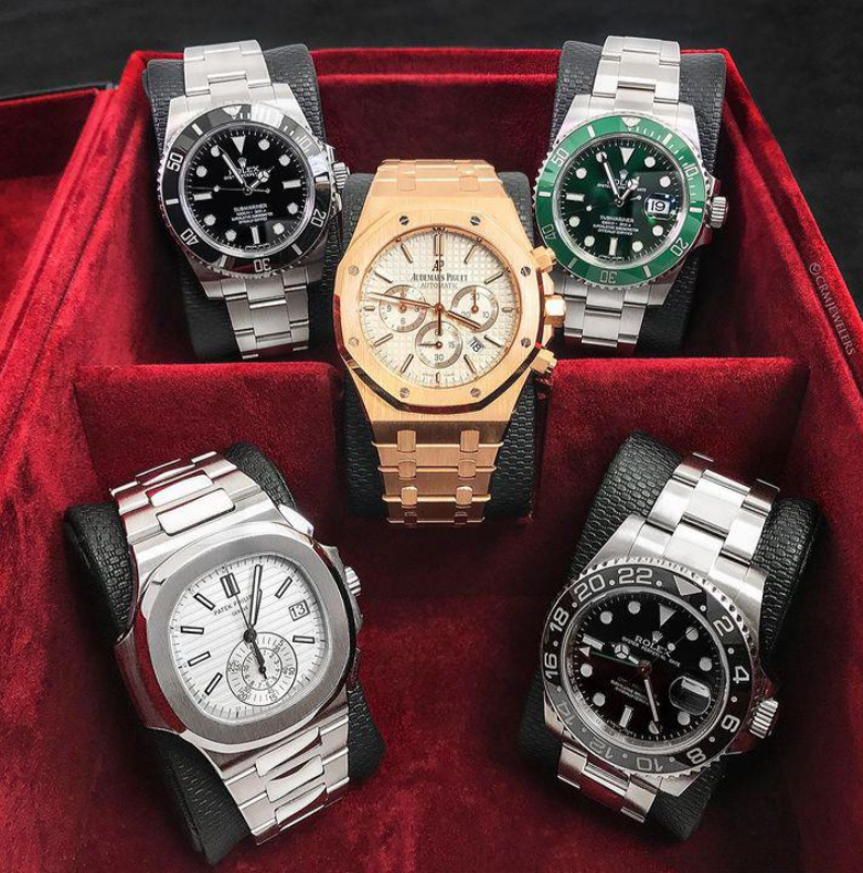 Five luxury wristwatches, including silver, gold, and two-tone models, are displayed in a red velvet watch box. Perfect to complement elegant clothes, these Rolex, Audemars Piguet, and Patek Philippe watches rest on black cushions.