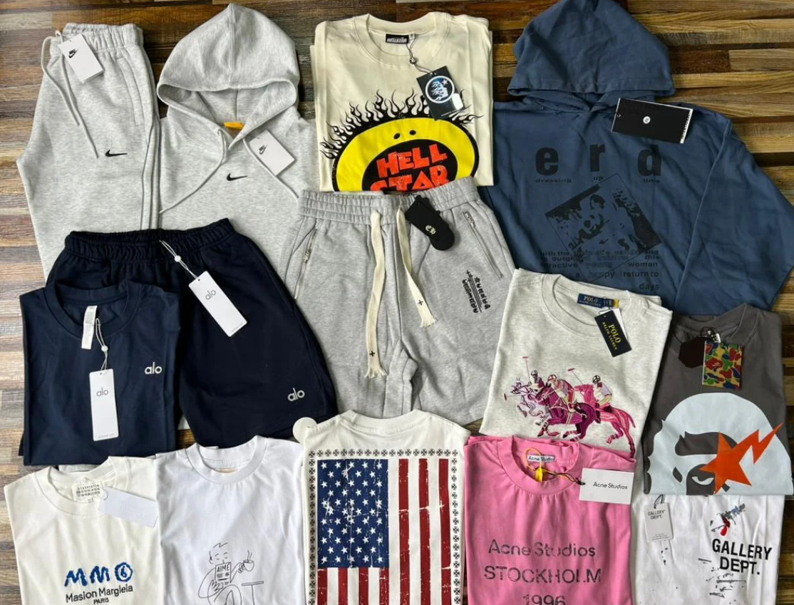 A collection of various branded clothes, including t-shirts, hoodies, shorts, and sweatpants, neatly arranged on a wooden floor. The shirts feature assorted graphics and logos, with tags visible on many items.