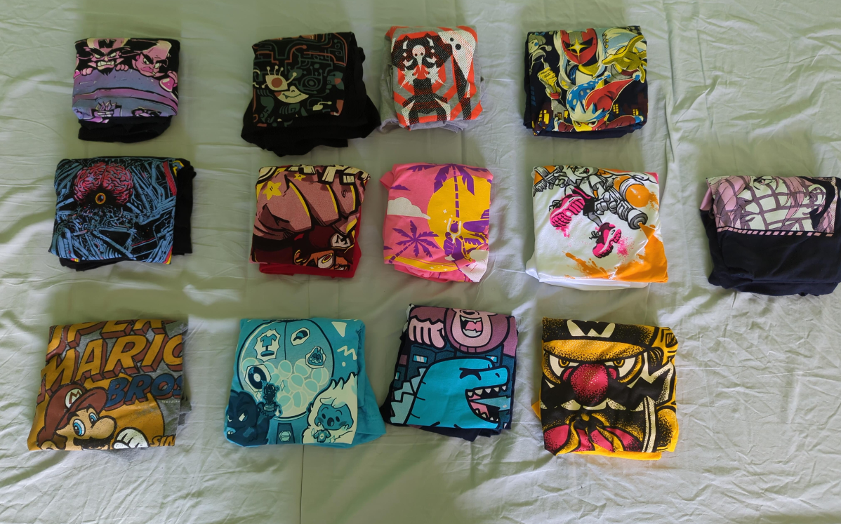 Twelve folded graphic t-shirts, perfect for anyone who loves fun clothes, feature colorful cartoon and video game designs. Arranged in three rows on a white bedsheet, each shirt showcases a unique character or themed illustration.