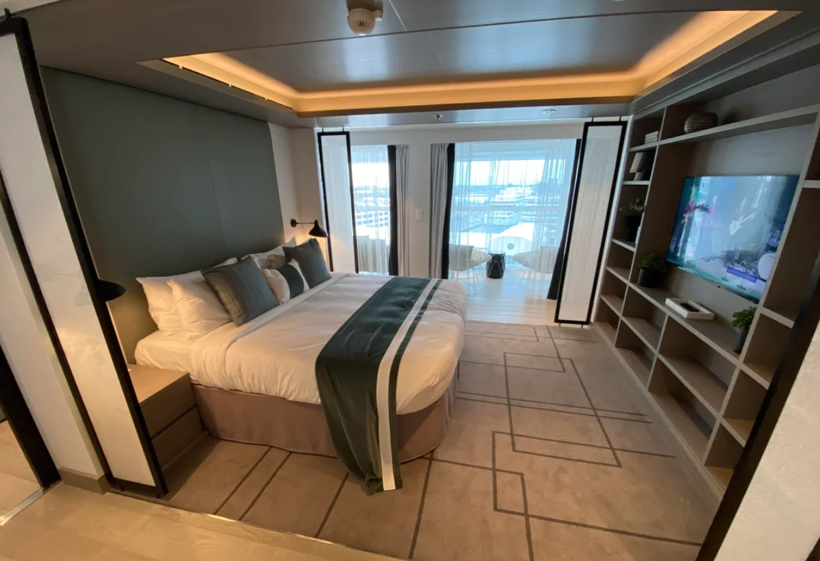 Modern hotel room with a large bed, stylish pillows, and a TV on a built-in shelf. Floor-to-ceiling windows with sheer curtains open to a balcony and waterfront view—perfect for relaxing or trying out your favorite travel hacks.