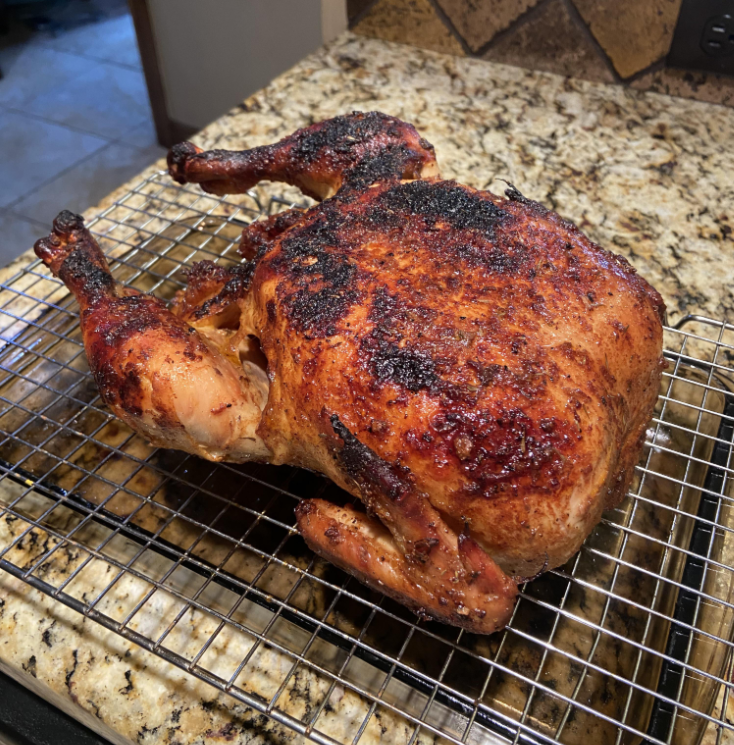 A whole roasted chicken with crispy, browned skin sits on a wire rack over a baking sheet, surrounded by fresh grocery items on a granite countertop.