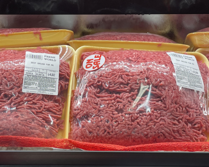 Packages of ground beef, wrapped in plastic, are displayed among other grocery items on a refrigerated store shelf. One package features a red "ON SALE" sticker, while the labels show product details and prices.