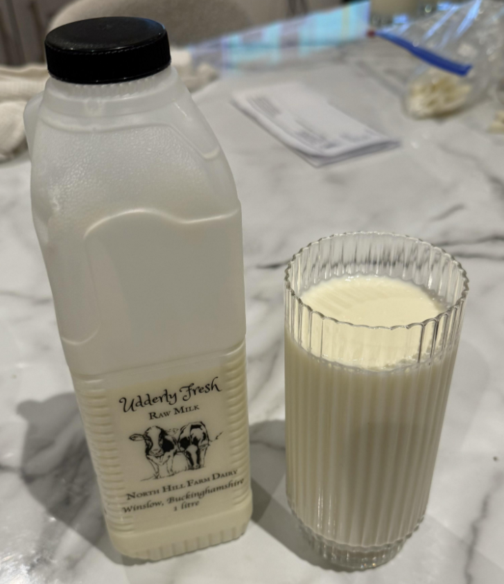 A carton labeled “Udderly Fresh Raw Milk” stands next to a tall, clear glass filled with milk on a marble countertop, surrounded by grocery items. A piece of mail and other items are visible in the blurred background.