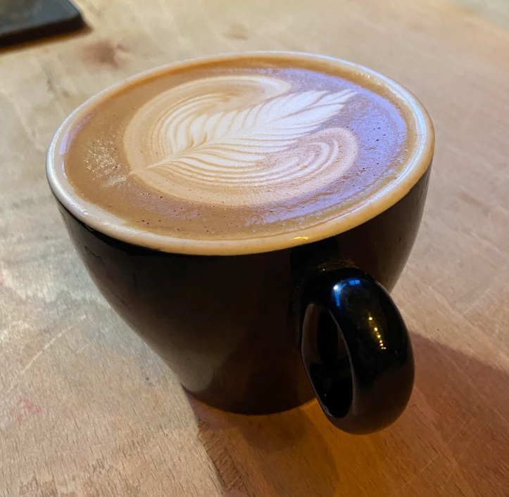 A black ceramic cup filled with coffee sits on a wooden table. The coffee, made from freshly ground grocery items, features a leaf-shaped latte art design on its creamy surface.