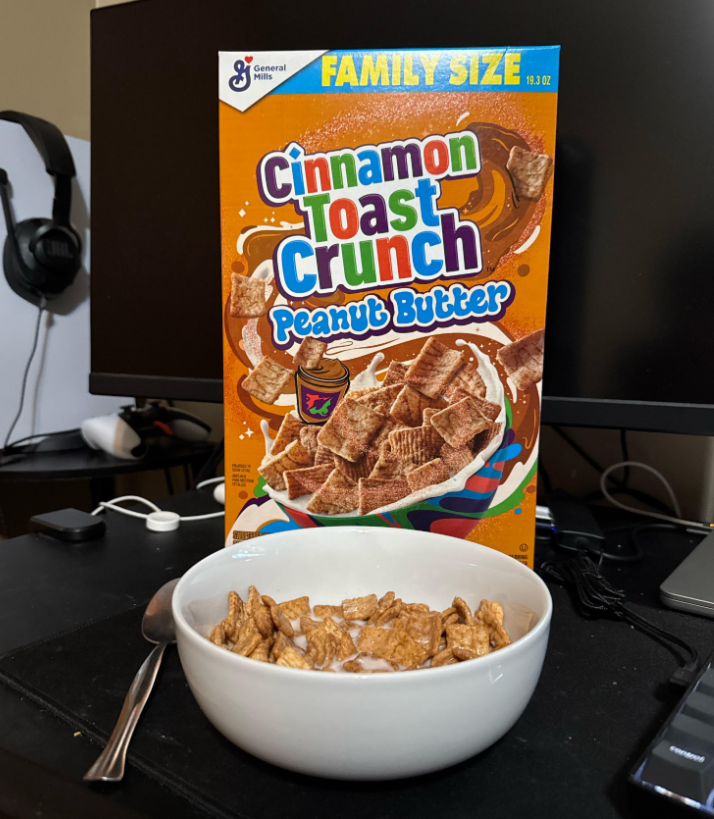 A bowl of Cinnamon Toast Crunch Peanut Butter cereal with milk sits in front of a "Family Size" box, surrounded by grocery items on the table, with a spoon beside it and a computer setup in the background.