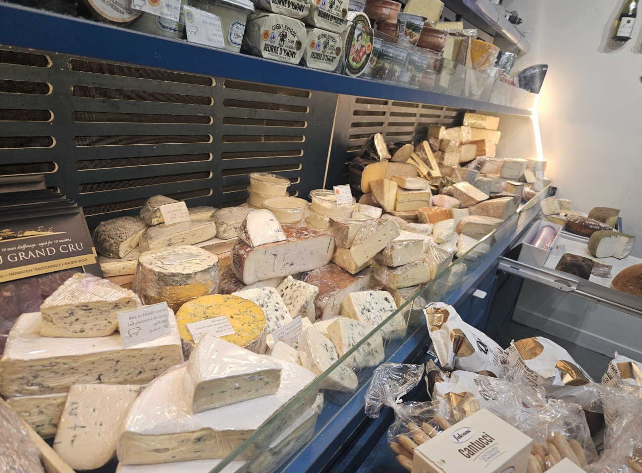 A display case filled with a wide variety of cheeses—including blocks, wedges, and rounds in different colors and textures—sits alongside labeled grocery items like packaged breadsticks in the foreground.