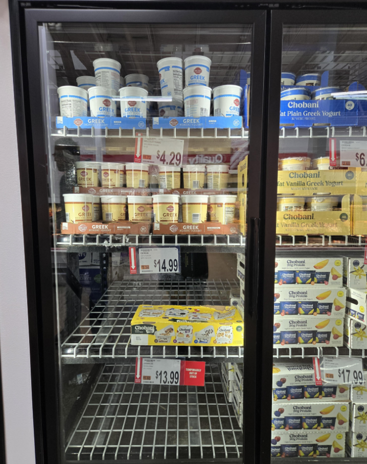 Refrigerated display case with various brands of Greek yogurt, including plain and vanilla flavors, in plastic tubs on shelves alongside other grocery items. Price tags read $4.29 and $14.99, with yogurt multipack boxes on the lower shelves.