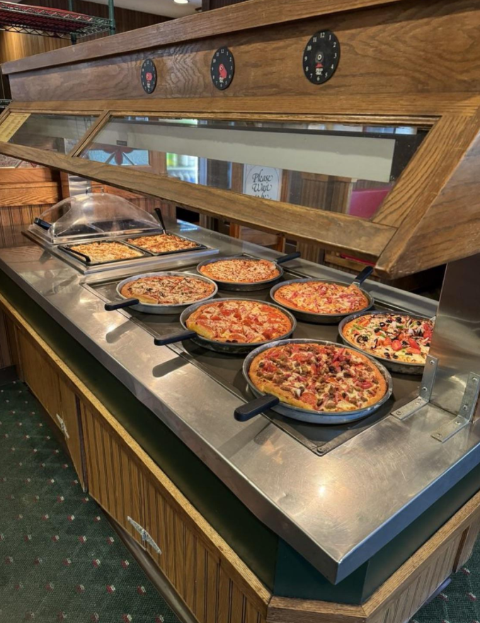 A pizza buffet with eight different pizzas in pans on a metal counter under a glass sneeze guard, offering a variety of toppings in a restaurant setting.