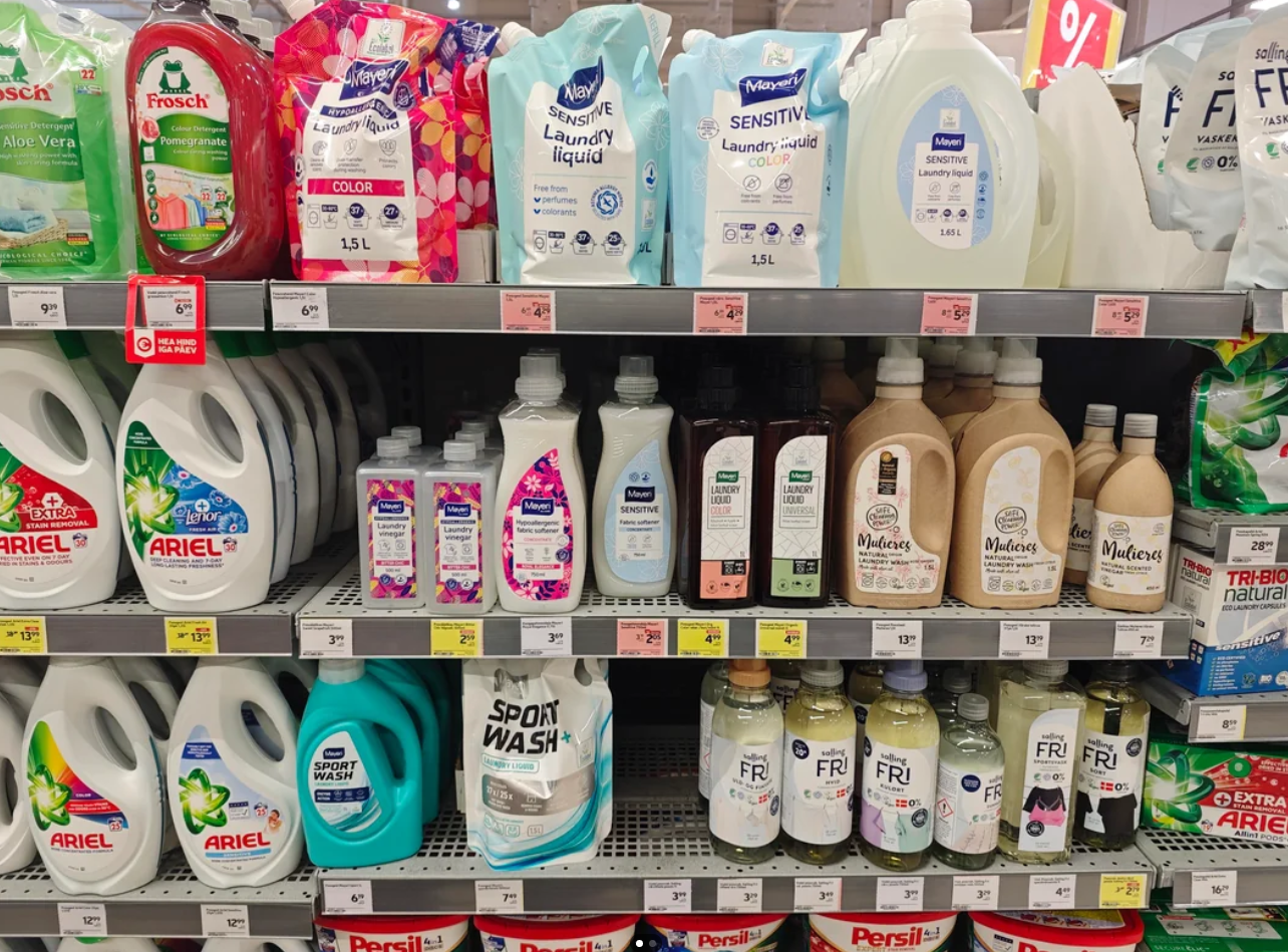 Bottles of various laundry detergents, displayed among other grocery items, line supermarket shelves. Price tags sit below each product, with large white bottles alongside smaller ones featuring colorful labels and brands.