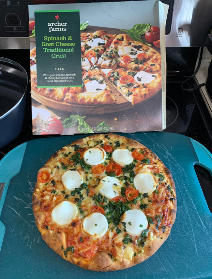 A baked spinach and goat cheese pizza, a standout among grocery items, sits on a blue cutting board in front of its Archer Farms box, which shows the same pizza topped with goat cheese rounds, spinach, and cherry tomatoes.
