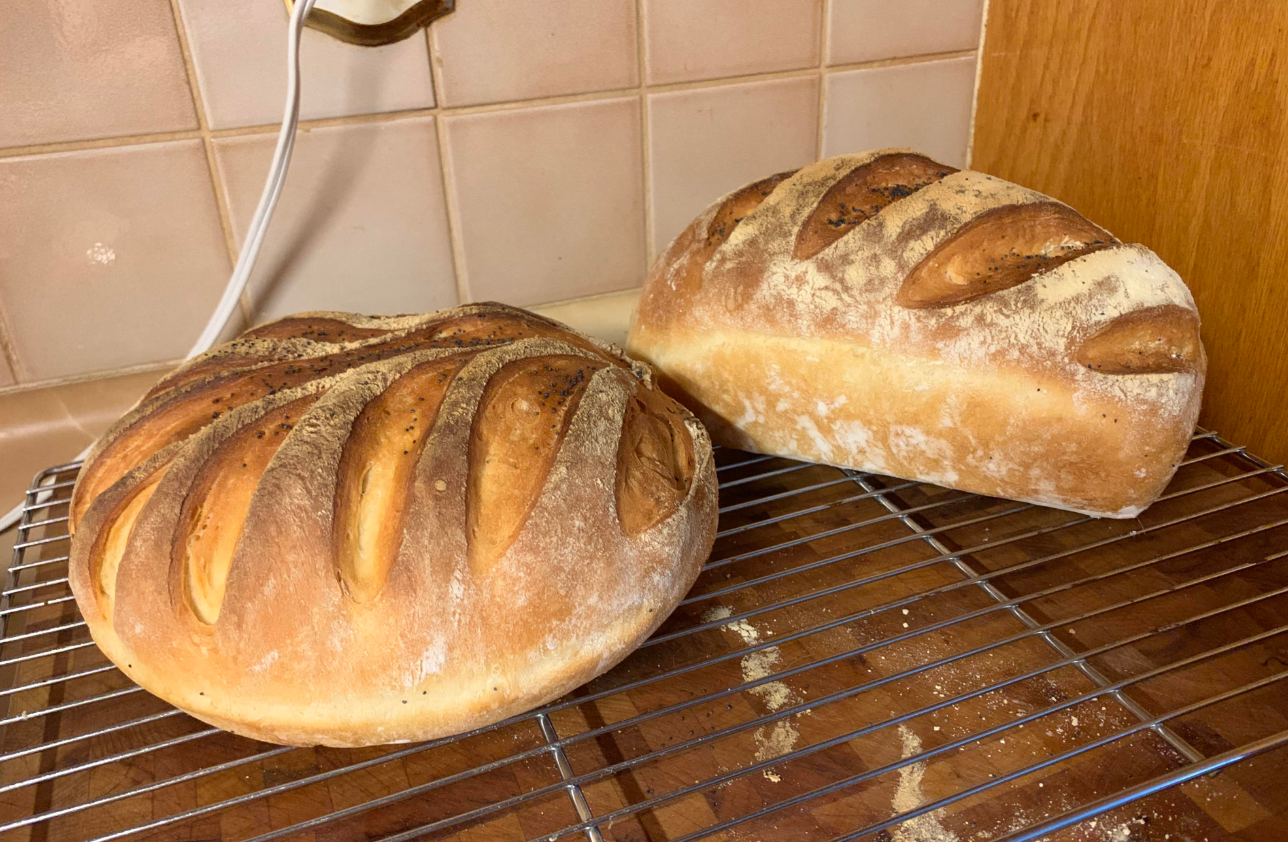 Two round loaves of freshly baked bread, classic grocery items with decorative slashes on top, cool on a wire rack in a kitchen with tiled walls and a wooden cabinet in the background.