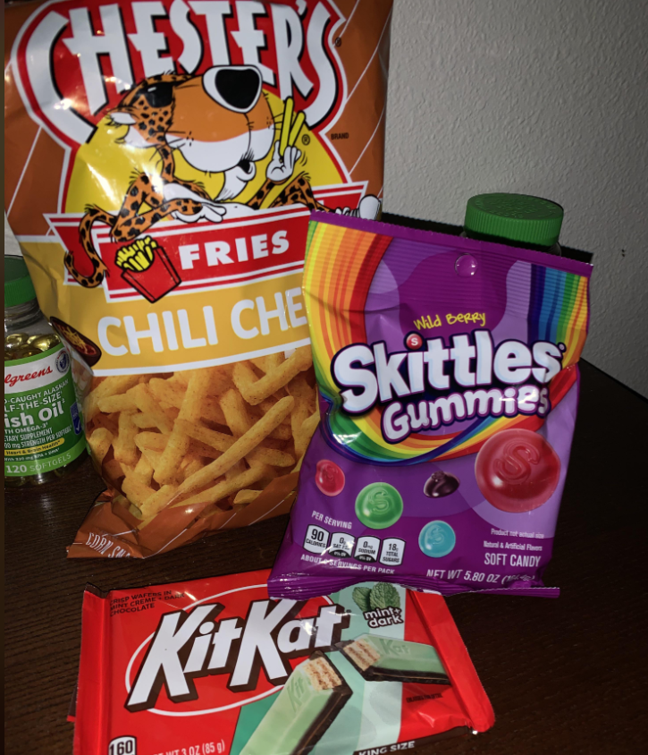 A selection of grocery items, including Chester's Fries Chili Cheese, Wild Berry Skittles Gummies, and a mint and dark chocolate Kit Kat bar, is arranged on a dark surface beside a bottle of green tea.