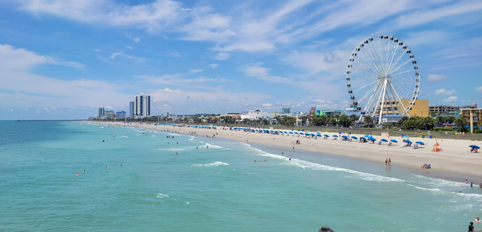 A sunny beach destination with turquoise waves, people swimming, and rows of blue umbrellas along the sand. Tall buildings and a large white Ferris wheel rise in the background under a partly cloudy sky.