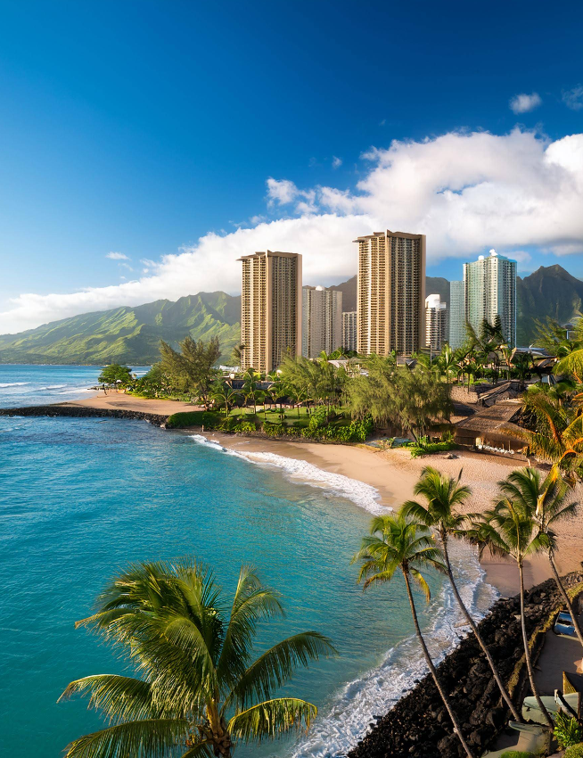 A tropical beach destination with turquoise water, palm trees, and sandy shore, backed by tall modern buildings and lush green mountains under a bright blue sky with scattered clouds.