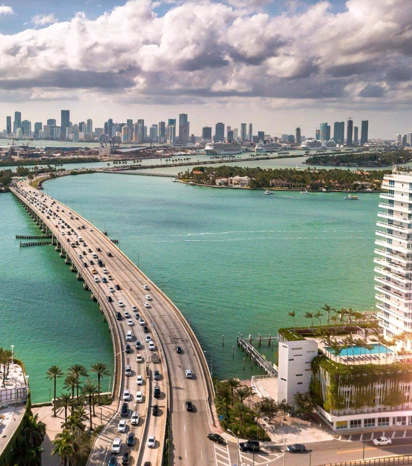 Aerial view of a busy causeway with cars crossing turquoise water toward a city skyline under partly cloudy skies, with modern buildings, palm trees, and small islands—perfect for exploring nearby beach destinations in the bay.