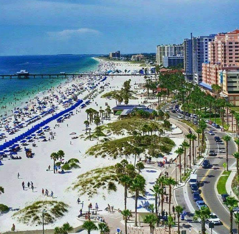 Aerial view of a popular beach destination with white sand, blue umbrellas, people sunbathing and swimming, palm trees, a curved road, and tall hotels and buildings lining the shore under a clear sky.
