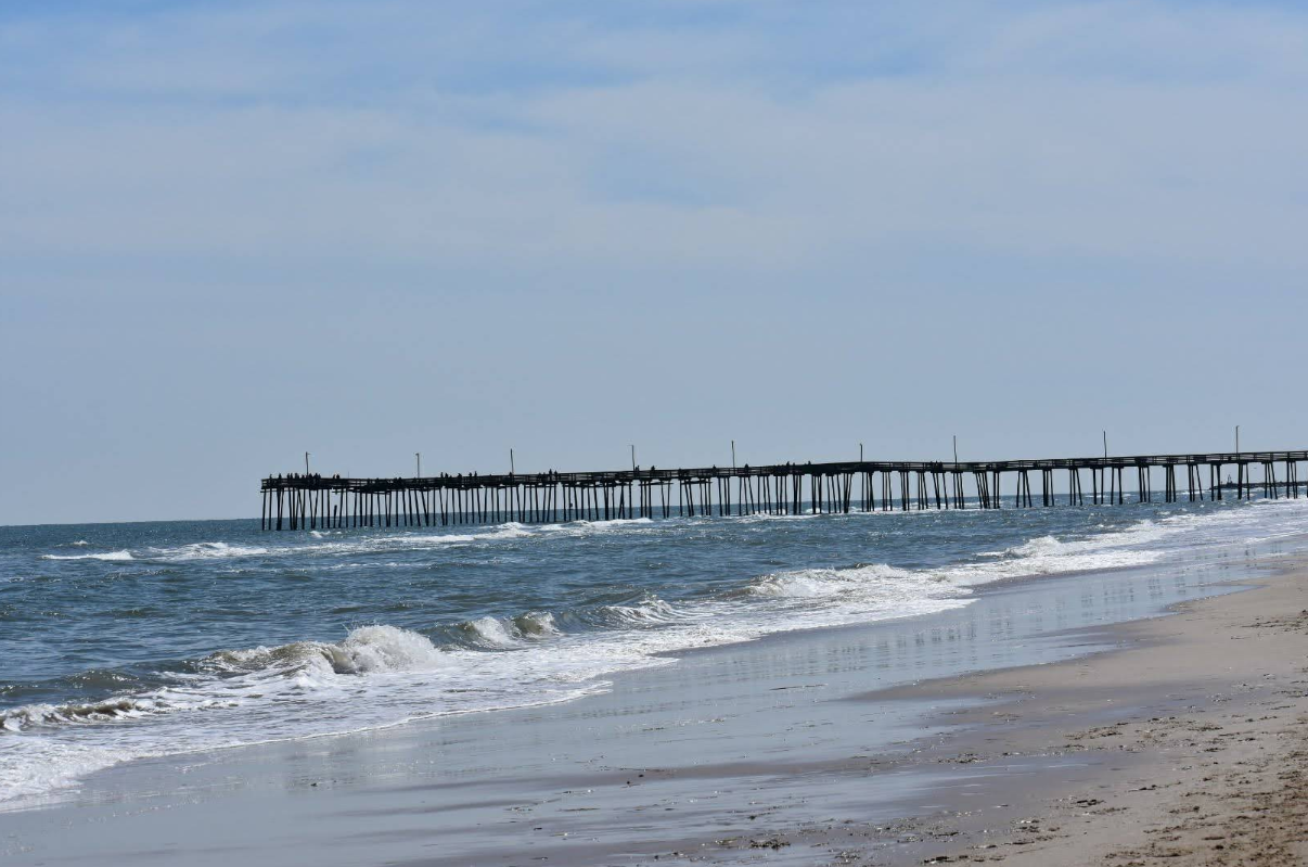 A long wooden pier stretches out over the ocean as gentle waves roll onto a sandy beach, capturing the serene beauty of sought-after beach destinations under a partly cloudy sky.