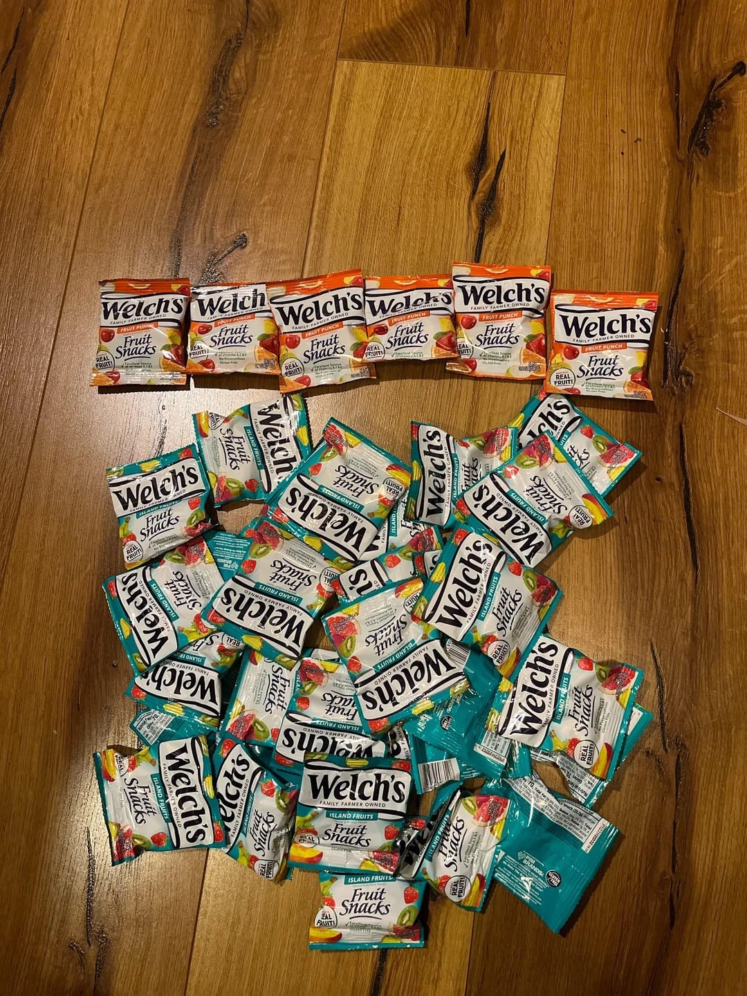 Twenty Welch’s Fruit Snacks packets, popular Walmart products, are scattered on a wooden floor—five orange tropical packets neatly in a row at the top and fifteen mixed fruit packets below in a loose pile.