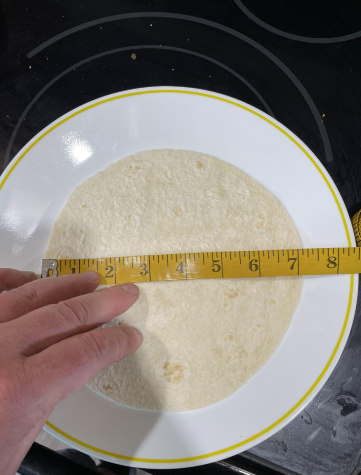 A hand holds a measuring tape across a flour tortilla on a white plate with a yellow rim, showing the tortilla measures about 7 inches in diameter. The plate is on a black stovetop.