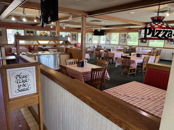 A cozy pizza restaurant dining area with wooden chairs, red and white checkered tablecloths, a salad bar, and a large hanging light that says "Pizza." A sign reads, "Please wait to be seated.