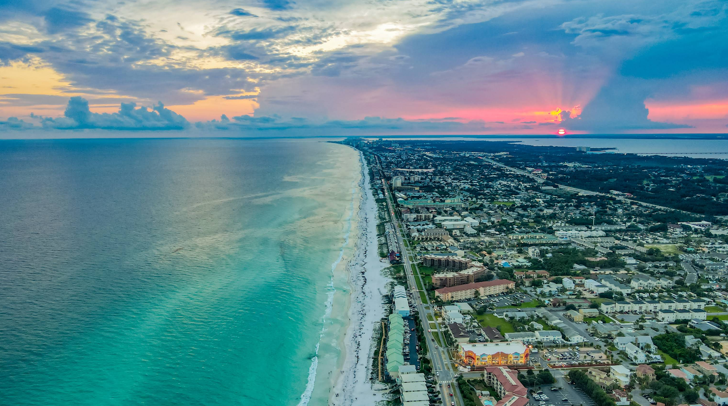 Aerial view of a coastal city at sunset, with turquoise ocean waves, a sandy beach, and buildings lined along a road. Perfect for beach destinations, the sky glows with colorful light as the sun sets near the horizon behind partly cloudy skies.