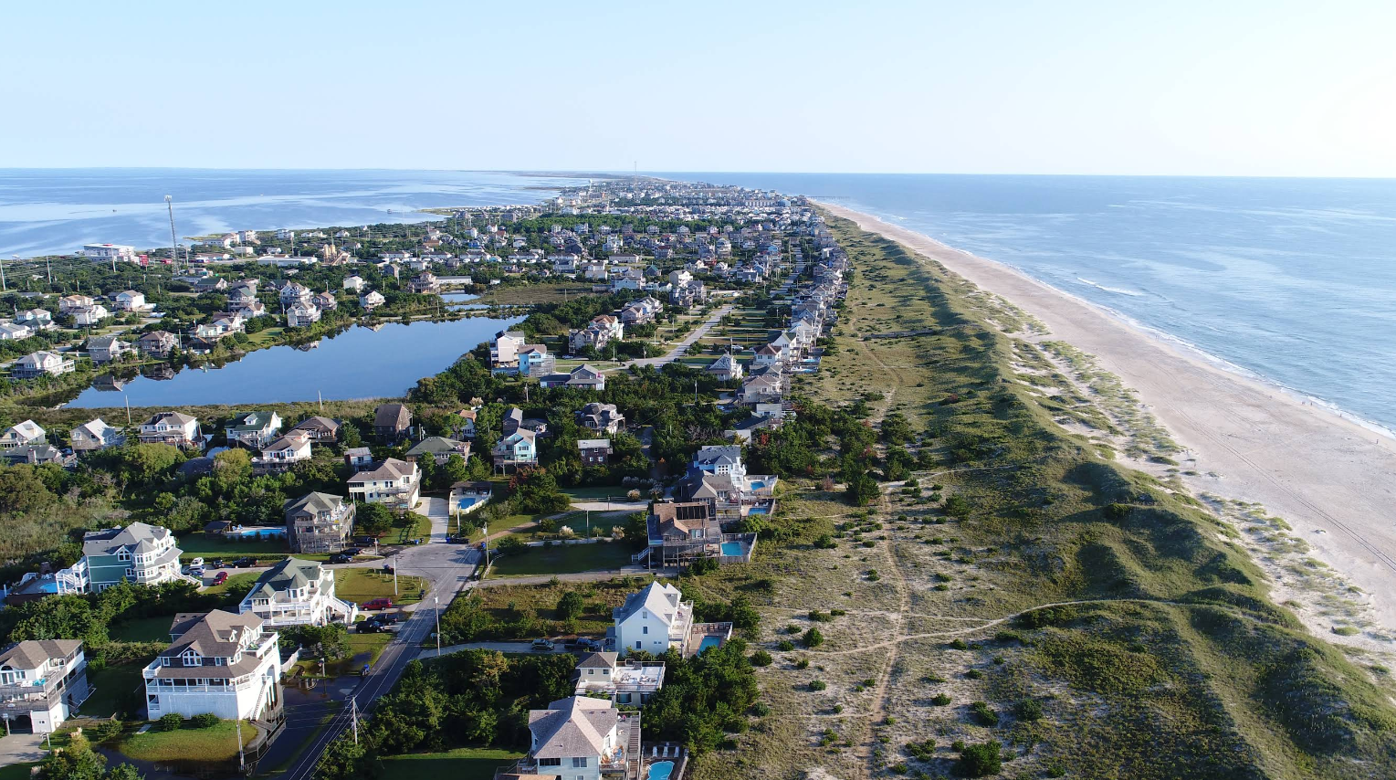 Aerial view of a coastal town, one of the top beach destinations, with houses along a narrow strip of land bordered by the ocean and a lake, featuring a sandy beach and grassy dunes running parallel to the shoreline.