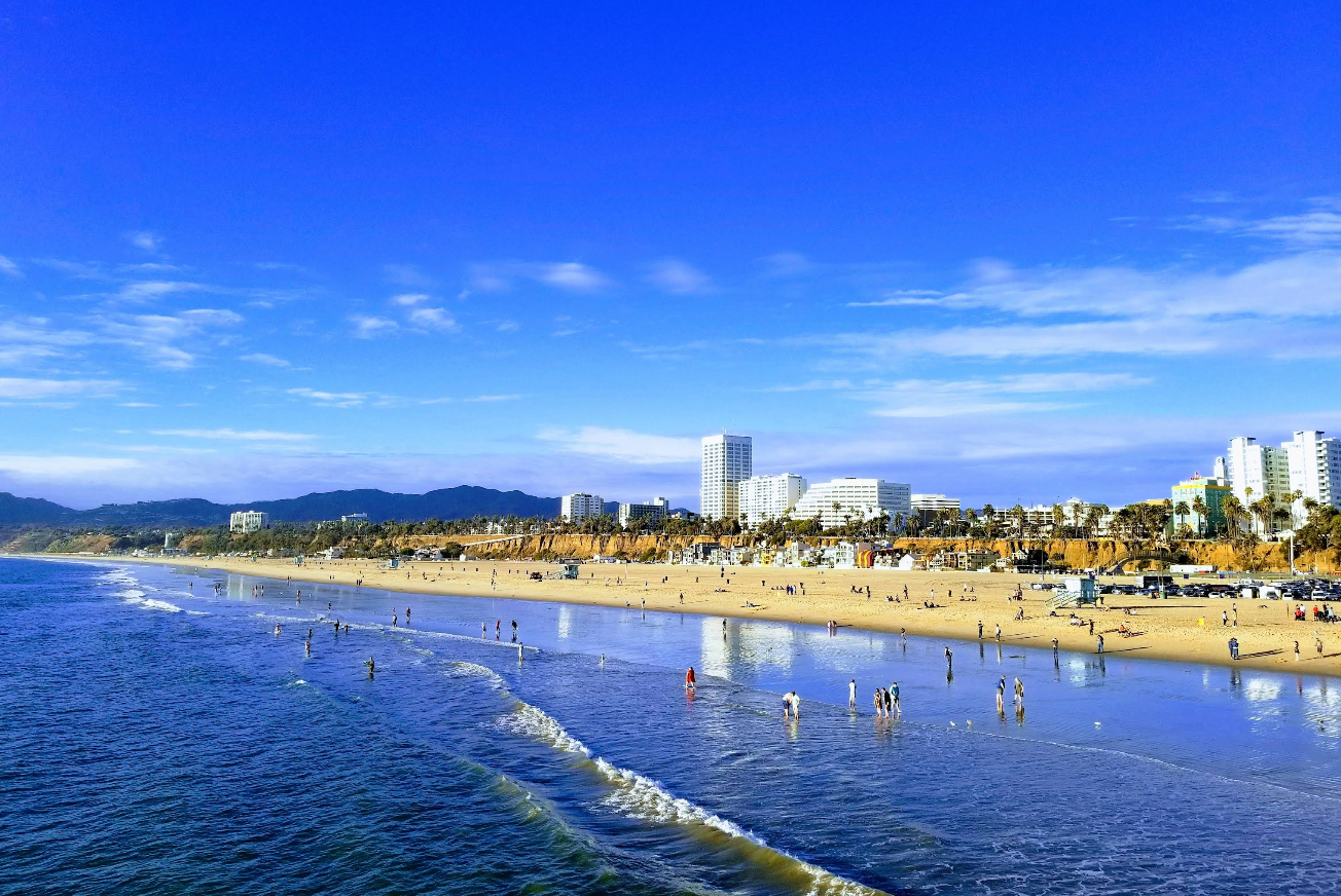 A wide sandy beach with people walking along the shore and wading in the water. In the background, tall buildings, palm trees, and distant mountains create a stunning scene typical of top beach destinations under a bright blue sky.