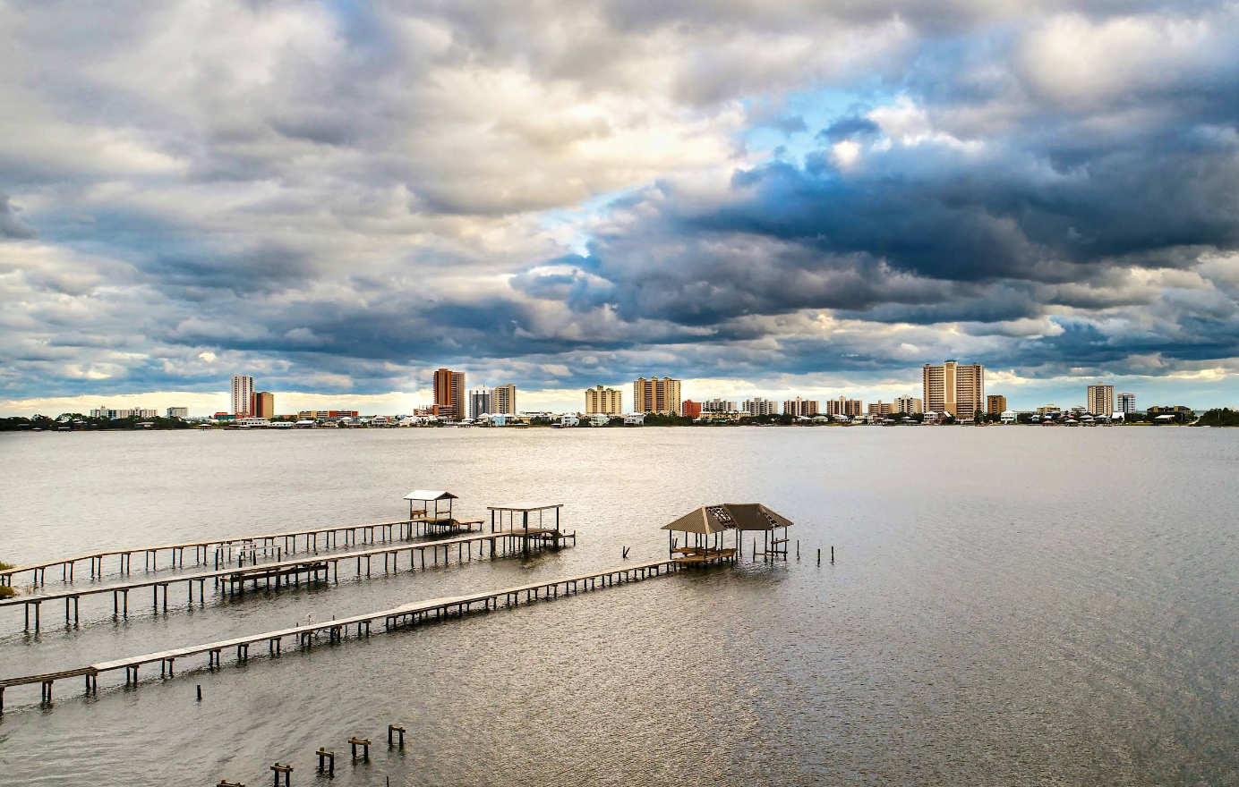 A view of a bay with wooden docks extending into the water, under a dramatic cloudy sky. High-rise buildings line the distant shore, highlighting one of the region's scenic beach destinations.