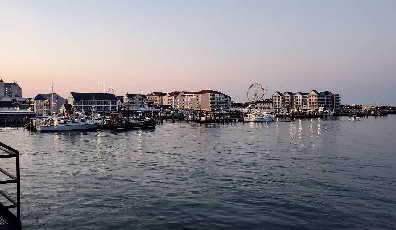 A calm harbor at dusk with boats docked near a waterfront lined with hotels and buildings. Lights reflect on the water, and a Ferris wheel rises in the distance, creating an inviting scene for those seeking serene beach destinations.