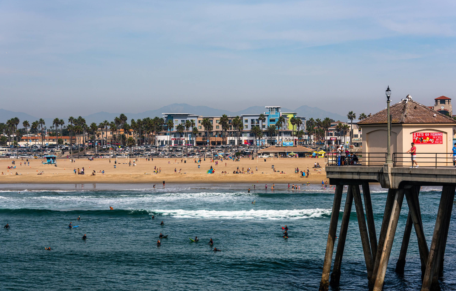 A lively beach scene at one of the top beach destinations, with people on the sand and surfers in the ocean, viewed from a pier with a red sign. Buildings and palm trees line the shore under a hazy sky.