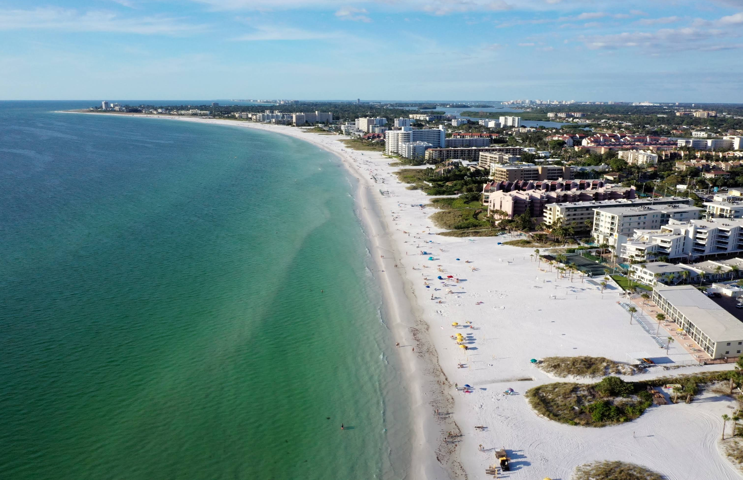 Aerial view of a top beach destination: white sandy shoreline curves along city hotels and condos, with turquoise water, scattered people, umbrellas, and greenery under a partly cloudy sky.
