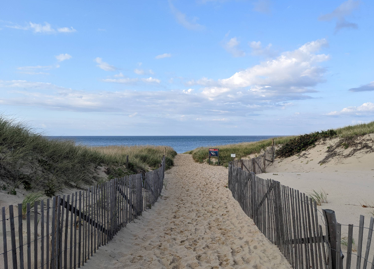 A sandy path lined with wooden fences leads through grassy dunes toward the ocean under a blue sky with scattered clouds—a classic scene among top beach destinations. A small sign is visible near the path before the beach.