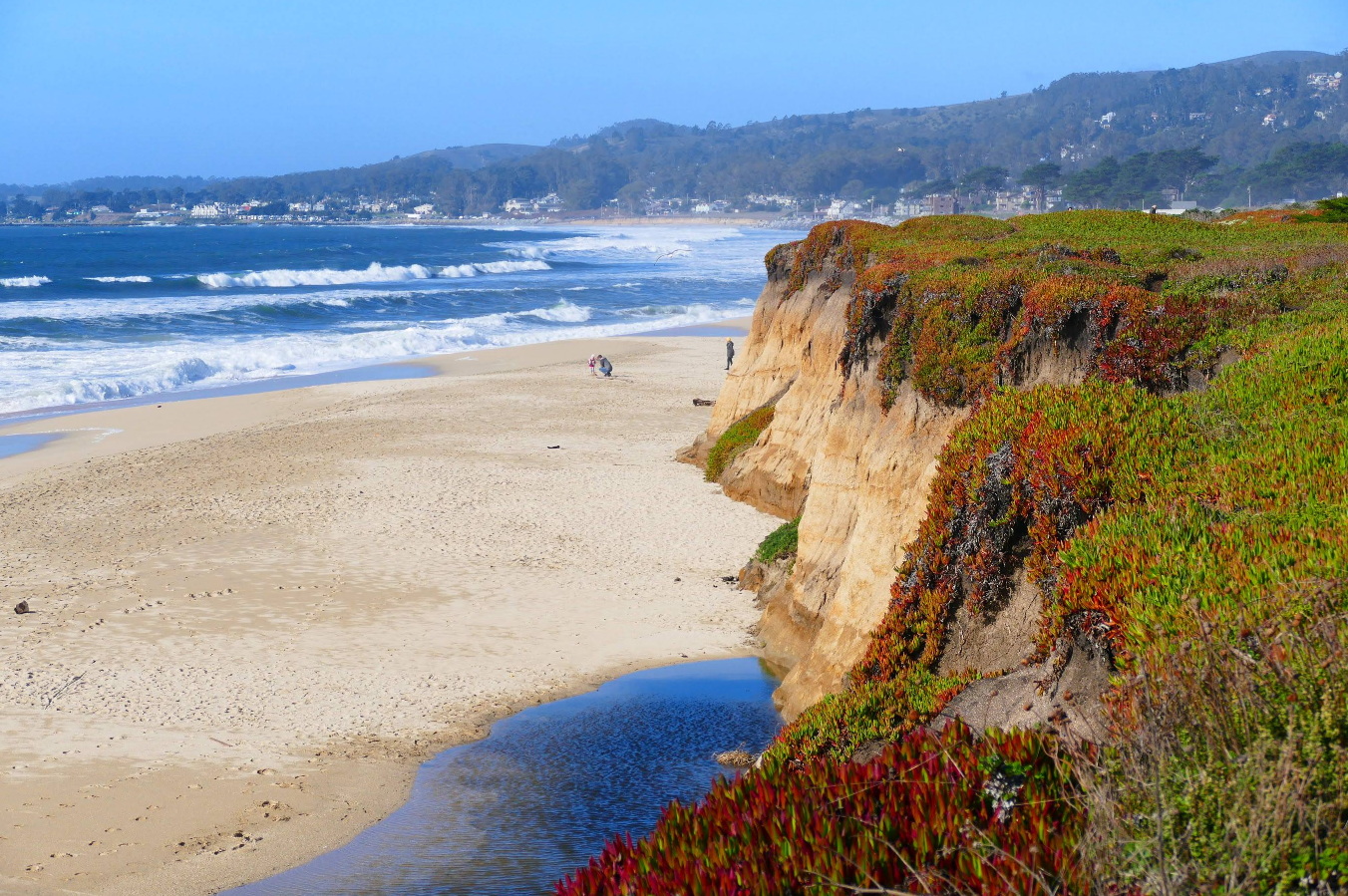 A sandy beach destination bordered by rugged cliffs with green and red vegetation, ocean waves, and distant hills under a clear blue sky. Two people walk along the shore near the water.