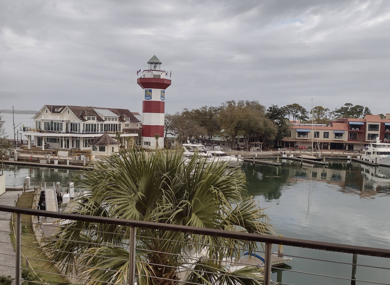 A marina with boats docked, a red-and-white striped lighthouse, palm trees, and waterfront buildings under a cloudy sky—one of the charming beach destinations perfect for relaxation and scenic views.