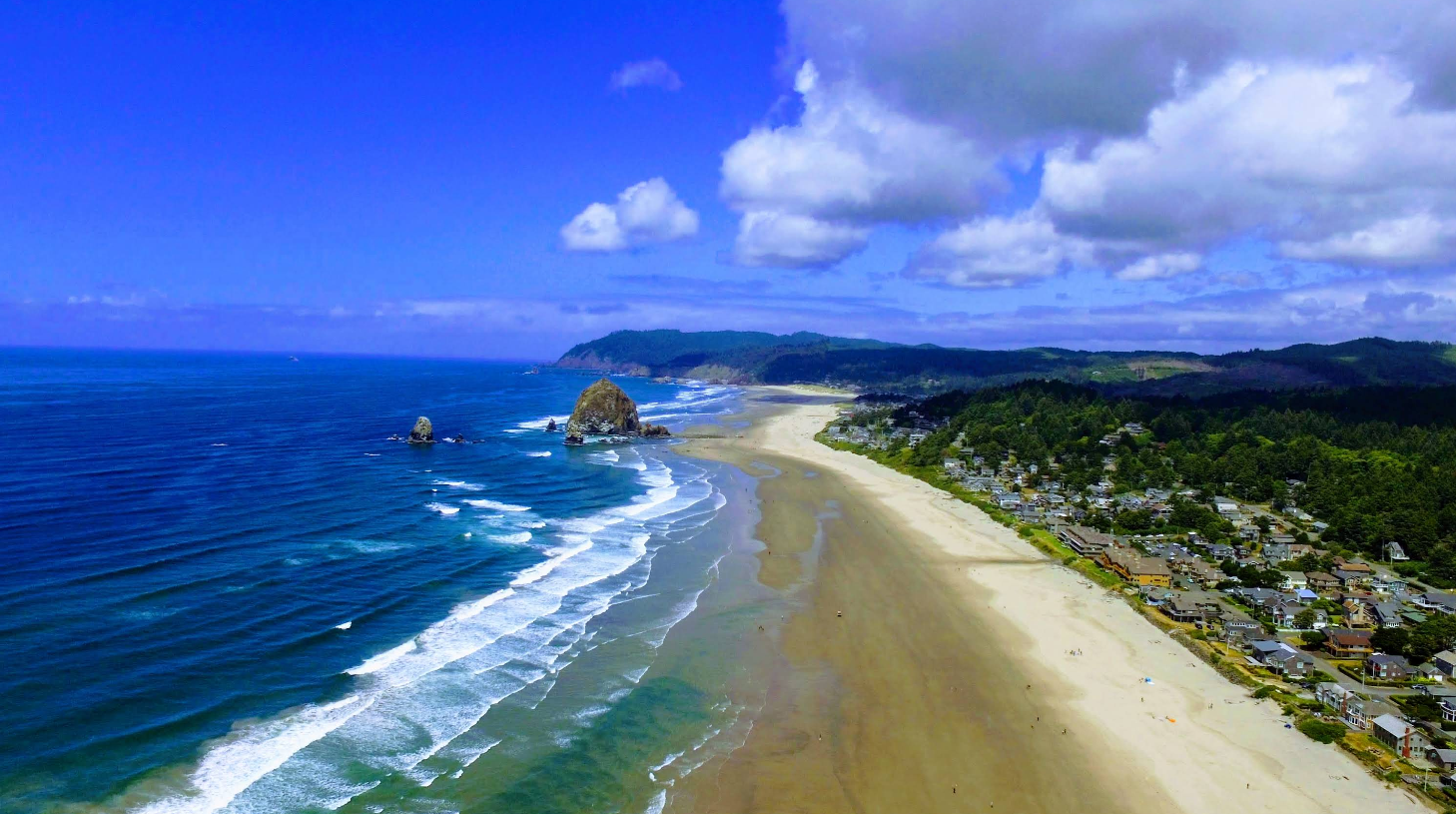 Aerial view of a sandy beach with waves rolling in, rocky formation in the water, green forested hills, and a small coastal town—an ideal scene for those seeking top beach destinations under a blue sky with scattered clouds.