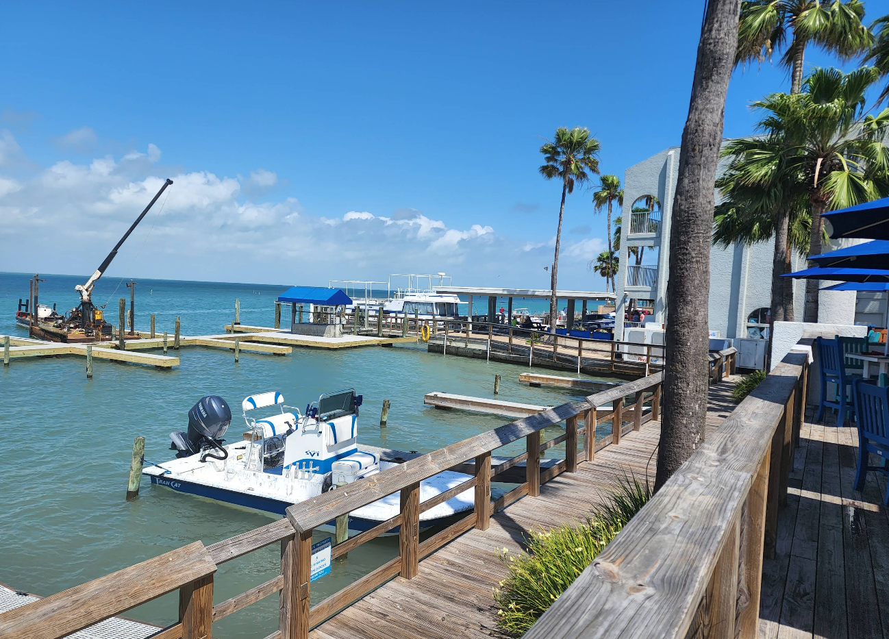 A marina with boat docks and a few boats on turquoise water, bordered by a wooden walkway with palm trees. Perfect for lovers of beach destinations, a crane works near the water and a modern building stands nearby under a bright blue sky.