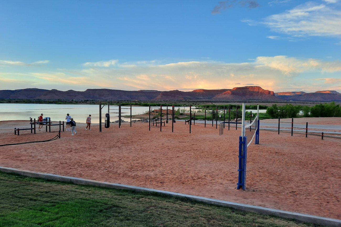 People play on a sandy volleyball court and use outdoor exercise equipment near a lake in one of the scenic tourist towns, with red rock mesas and a partly cloudy sky glowing at sunset.