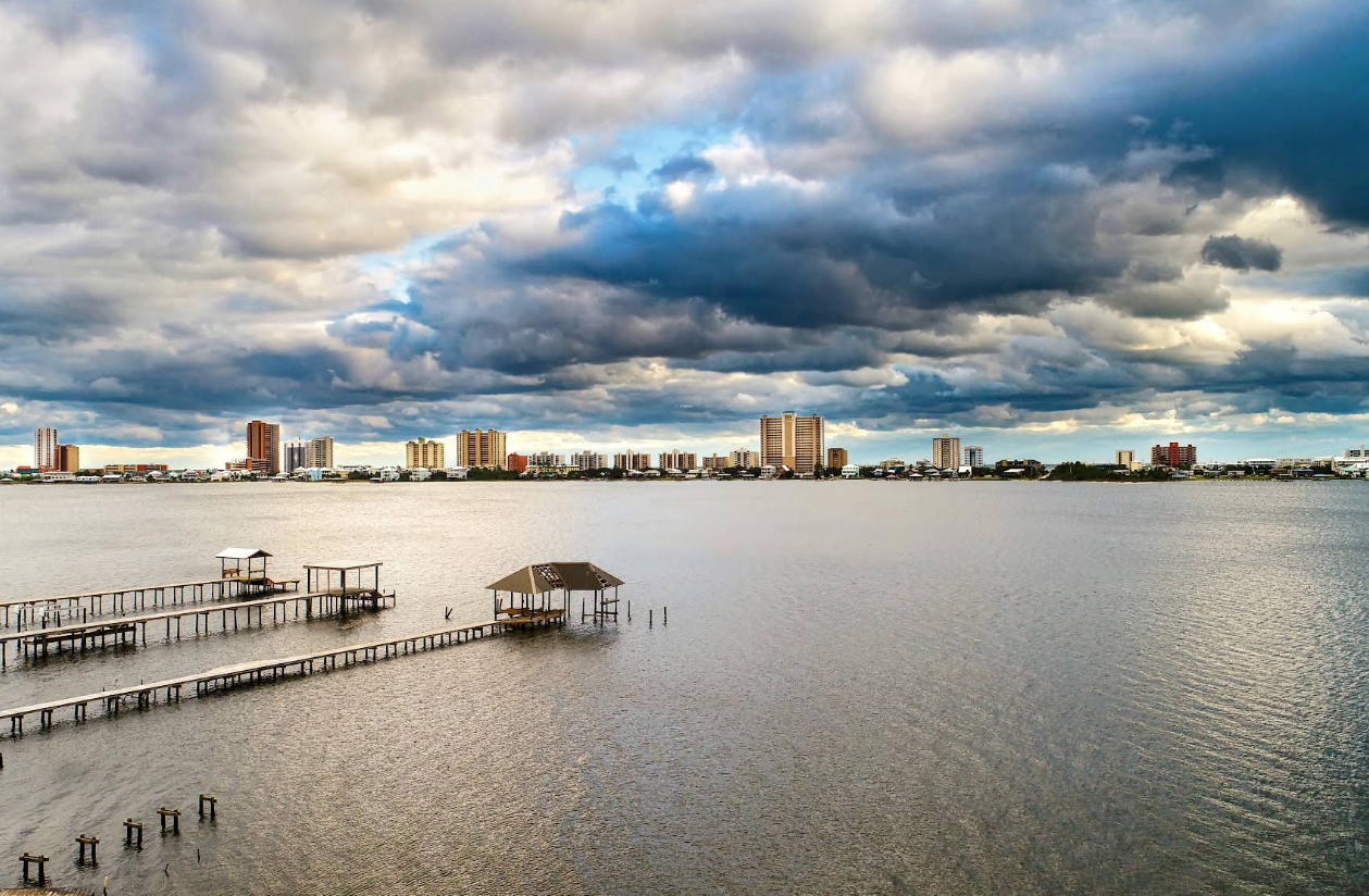 A wide river or bay with a wooden pier and small gazebo extends over the water; tall buildings line the distant shore, evoking the charm of tourist towns beneath a dramatic cloudy sky.