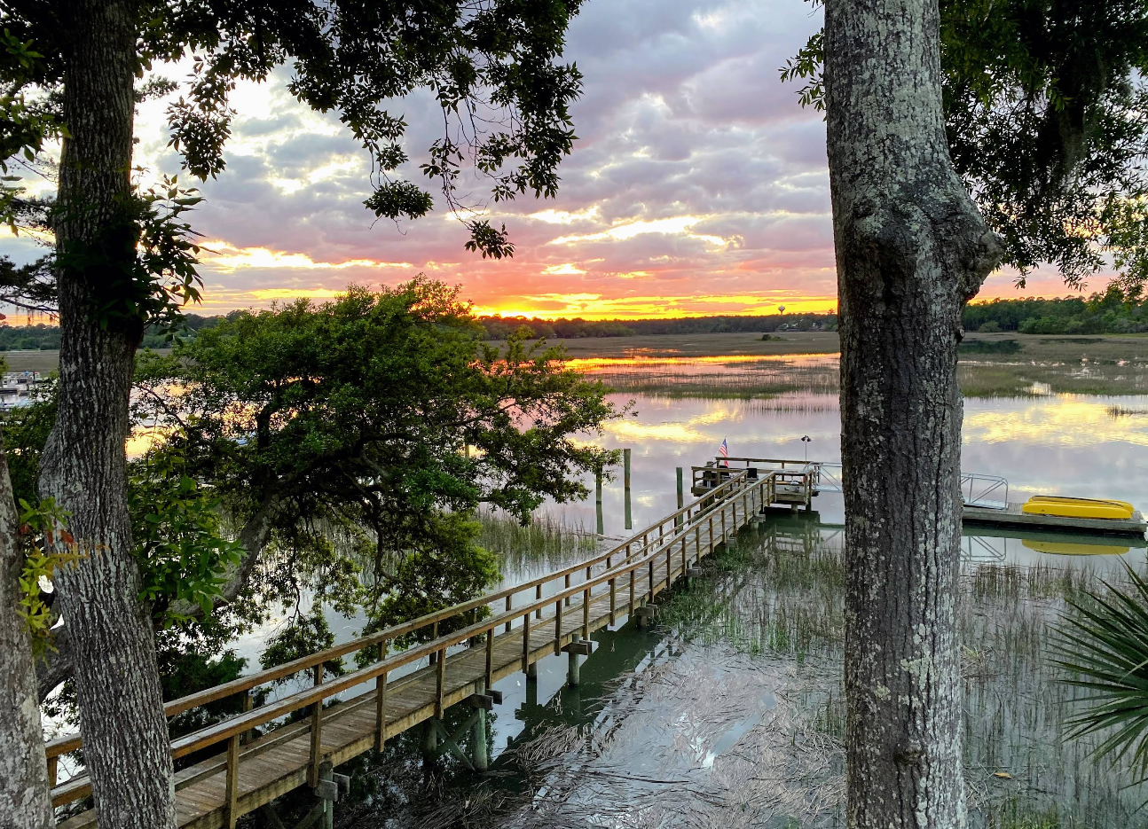 A wooden dock extends over calm water surrounded by trees and marshland at sunset, with vibrant orange and yellow hues reflecting on the water’s surface—a serene retreat often found in charming tourist towns. A boat is tied to the dock.