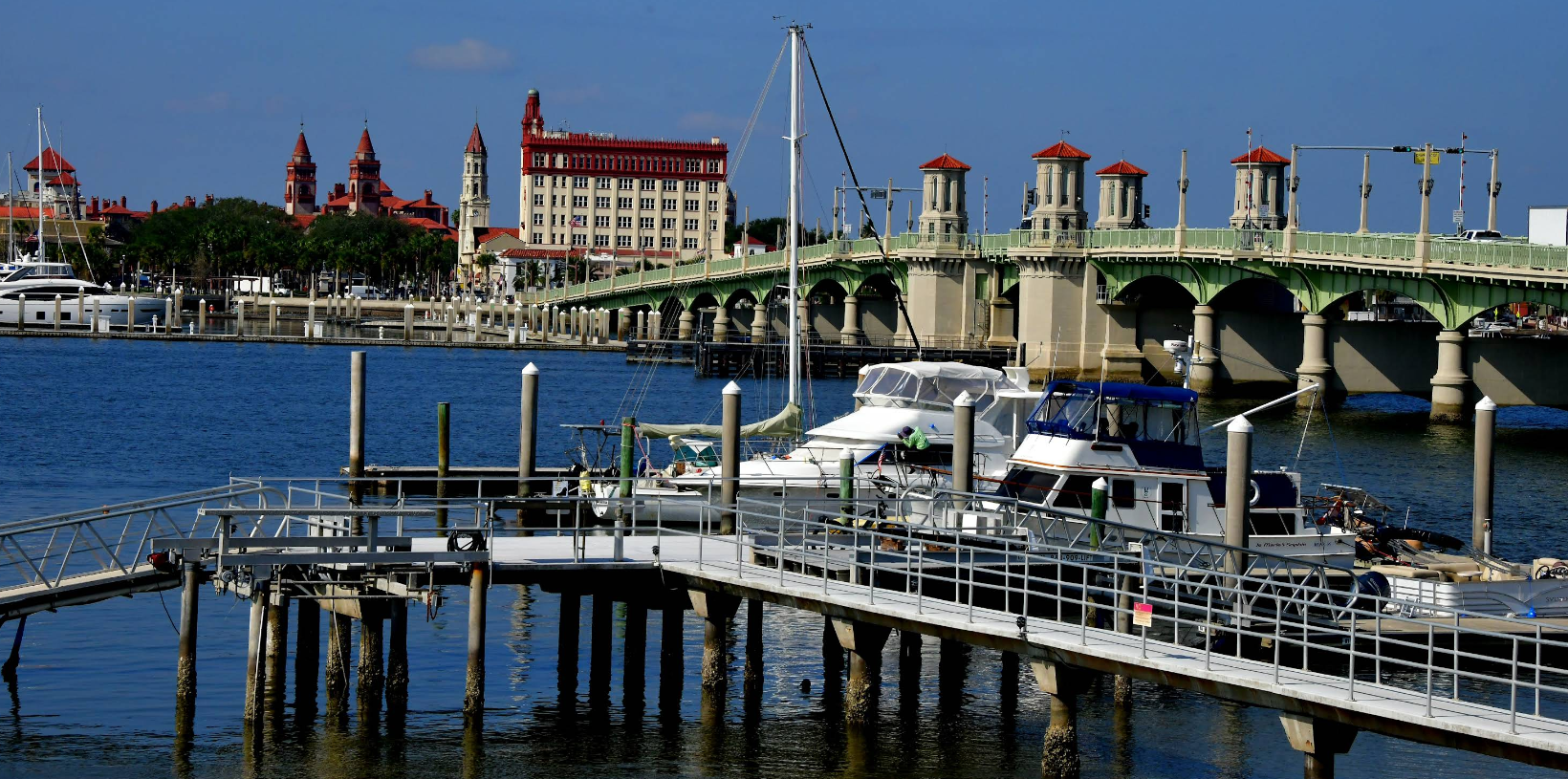 A marina with boats docked, a green bridge spanning the water, and historic buildings with red roofs evoke the charm of many tourist towns under a clear blue sky.