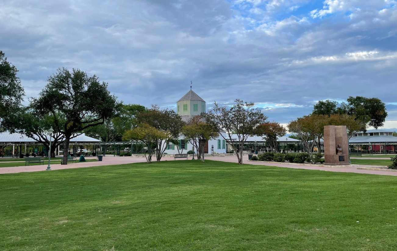 A wide grassy lawn with scattered trees, a central white building with a tower, and a tall stone monument under a partly cloudy sky evoke the charm often found in tourist towns.