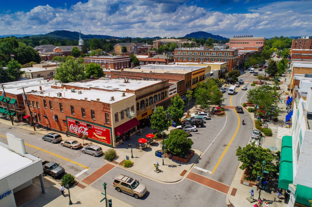 Aerial view of a tourist town’s vibrant main street lined with historic brick buildings, shops, parked cars, outdoor seating, and trees, with hills and a partly cloudy sky in the background.
