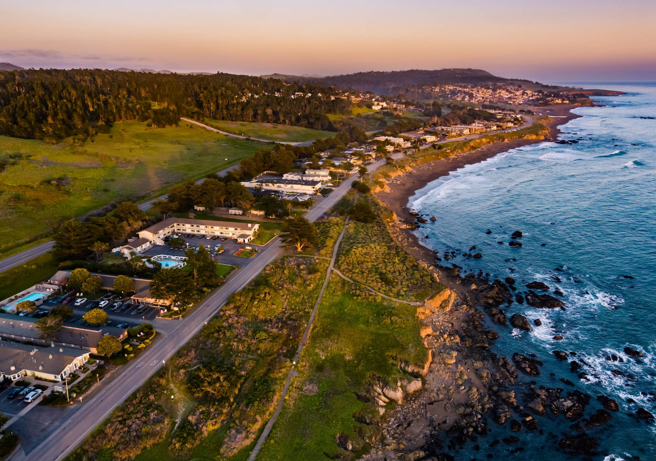 Aerial view of one of the scenic tourist towns at sunset, with buildings along a winding road, green hills, and rocky shoreline next to the ocean. The sky has a soft golden hue.
